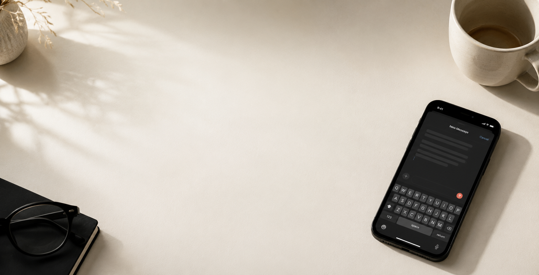 A black smartphone lying on a light beige desk next to a white coffee mug, with a notebook and glasses partially visible in the frame.